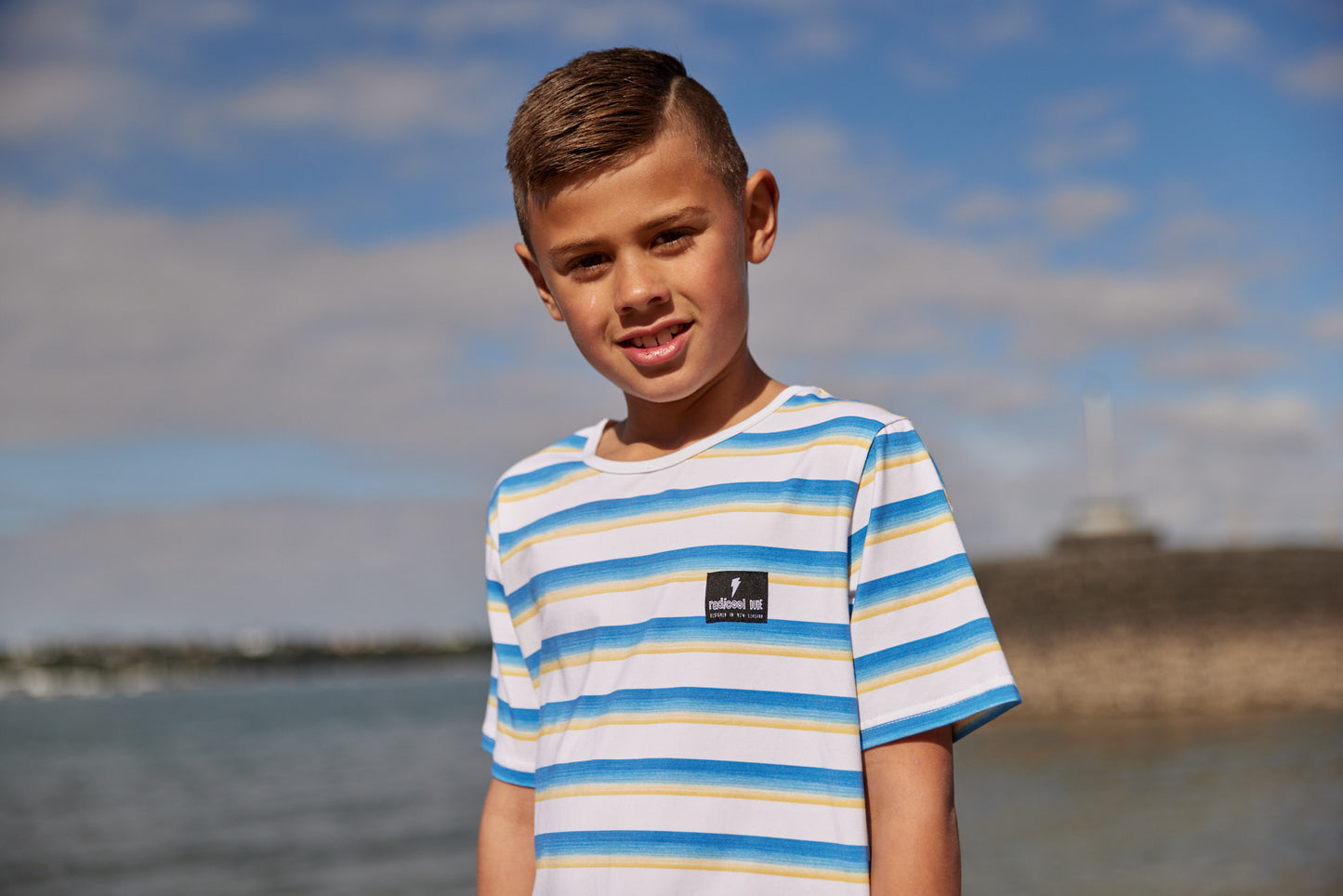 Radicool Kids beach stripe tee: Cotton / spandex tee in white with beach waves printed in horizontal stripes. Radicool Dude badge on chest. We love a stripe, even more when it’s the colours of the beach! Fit is true to size.